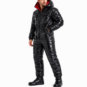 Op de afbeelding: Een zwarte, hoogglanzende, gewatteerde jumpsuit uit één stuk met een ritssluiting over de hele lengte aan de voorzijde, lange mouwen met elastische boorden en een capuchon met een felrode voering. Het kledingstuk heeft een getailleerde taille en bijpassende zwarte laarzen.