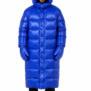 Puede incluir: Un abrigo largo de plumas en color azul brillante con diseño acolchado, una capucha grande y cierre frontal con botones a presión. El abrigo incluye manoplas azules a juego y se lleva sobre pantalones negros y botas de color negro.