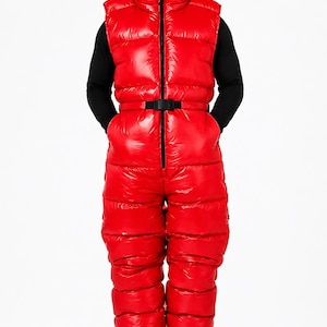 May include: A bright red, sleeveless, quilted puffer jumpsuit featuring a high collar, a full-length front zipper, and a black adjustable waist belt with a plastic buckle. The garment has a shiny, smooth finish and horizontal baffle construction.