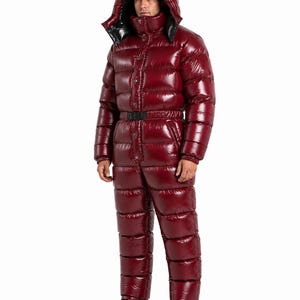 May include: A glossy, deep burgundy, one-piece quilted puffer jumpsuit featuring a large, insulated hood with a black interior lining, a front button closure, a black waist belt with a buckle, and long sleeves with elasticated cuffs.
