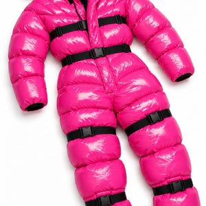 Op de afbeelding: Een felroze, hoogglanzende gewatteerde jumpsuit met capuchon, lange mouwen en een ritssluiting aan de voorzijde. Het kledingstuk is voorzien van meerdere zwarte, verstelbare banden en kunststof gespsluitingen over de borst, taille en benen.