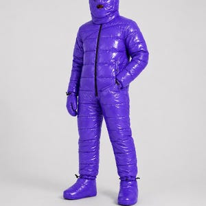 Pode incluir: Um macacão acolchoado de corpo inteiro em roxo brilhante com acabamento lustroso. A peça apresenta um capuz com abertura para o rosto, mangas compridas, luvas a combinar e botins integrados. Um fecho de correr preto no centro.
