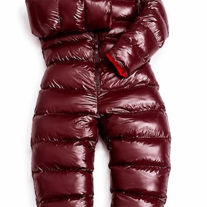 Op de afbeelding: Een glanzend, donkerbordeauxkleurig gewatteerd jumpsuit voor het hele lichaam met lange mouwen en een capuchon. De binnenvoering van de capuchon en de mouwboorden is felrood en vormt een contrast. Een centrale rits loopt over de voorkant.