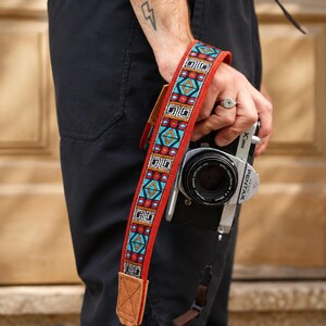 Puede incluir: Una correa de cámara con un patrón tejido vibrante en turquesa, rojo y negro, bordeada por tela roja y terminada con extremos de cuero beige. La correa está unida a una cámara Pentax plateada y negra con un objetivo circular y cuerpo negro.