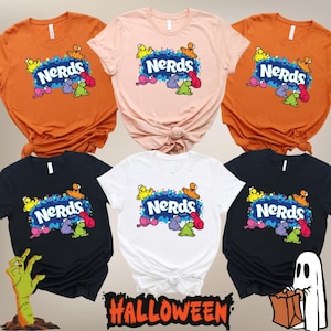 May include: A collection of five short-sleeved t-shirts in orange, light pink, black, and white colors. Each shirt features a central graphic of the Nerds candy logo with colorful cartoon characters surrounding the text on a blue splattered background.