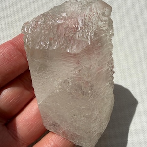 Puede incluir: Un espécimen de cristal de cuarzo transparente y en bruto con una textura superficial irregular y dentada. El mineral presenta facetas naturales e inclusiones internas, mostrando una estructura cristalina con bordes afilados e irregulares.