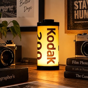 Może przedstawiać: Cylindryczna lampa w kształcie starej rolki filmu ze świecącym żółtym korpusem. Napis Kodak 200 jest nadrukowany na czarno z boku. Lampa posiada czarne okrągłe nakładki na górze i na dole oraz emituje ciepłe światło ze swojego wnętrza.