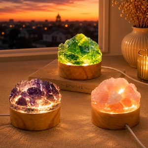 May include: Three circular lamps featuring raw crystal clusters in green, purple, and pink, each mounted on a cylindrical wooden base. The lamps are illuminated from within, casting a warm glow through the translucent mineral formations.