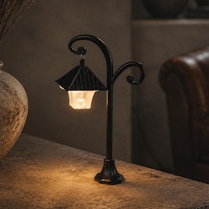 May include: A black metal miniature street lamp with a curved post and a decorative scroll arm. The lamp features a square, ridged roof and a translucent, glowing lantern housing that emits a warm, soft light from an internal light source.