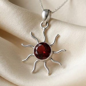 Puede incluir: Un collar de plata con un colgante en forma de sol que presenta una piedra granate roja oscura, redonda y facetada en el centro. El colgante tiene doce rayos curvos y ondulados que se extienden desde la piedra central, en una cadena fina.