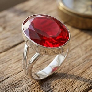 May include: A silver ring featuring a large, oval-cut, deep red gemstone set in a polished metal bezel. The band of the ring is split into two distinct sections that meet at the base, providing a structured and open design for the jewellery piece.