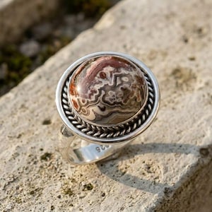 Puede incluir: Un anillo de plata con una piedra cabujón redonda y pulida que presenta intrincados patrones de remolinos en tonos marrón, crema y blanco. La piedra está engastada en un borde de metal trenzado y el aro es de plata lisa y pulida.