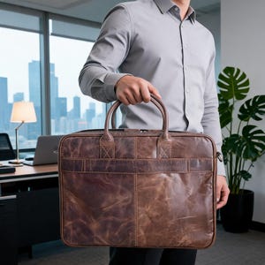 May include: A rectangular brown leather briefcase with a distressed finish, featuring two top handles, a horizontal stitched strap across the center, and a top zipper closure. The bag has visible grain texture and reinforced stitching at the handle bases.