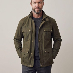 May include: An olive green field jacket featuring a high collar with a snap-button tab, a front zipper closure, and four large patch pockets with snap-button flaps. The jacket is worn over a dark gray crew-neck sweater and dark gray trousers.