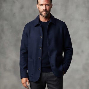Puede incluir: Una chaqueta azul marino con botones, cuello y dos bolsillos frontales de parche, sobre un suéter de cuello redondo azul oscuro y pantalones gris carbón. La chaqueta tiene cuatro botones visibles en la parte delantera y mangas largas.