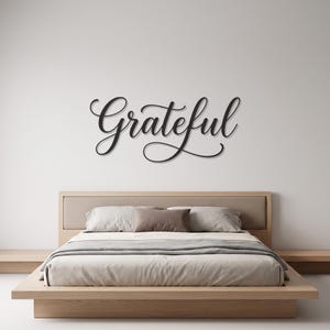 May include: A black metal wall sign featuring the word Grateful in a cursive script font. The lettering is mounted on a plain white wall above a low-profile wooden bed frame with light gray bedding and a single brown accent pillow.