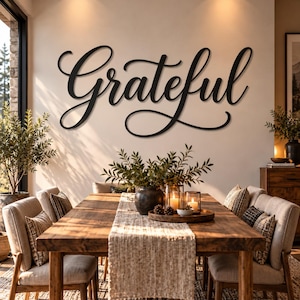May include: A black metal wall sign featuring the word Grateful in a cursive script font. The sign is mounted on a light-colored wall above a wooden dining table, which is set with a centerpiece of green foliage in a dark vase and candles.