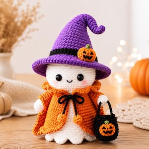 Può includere: Una bambola fantasma all'uncinetto bianca che indossa un cappello a punta viola con un piccolo dettaglio a forma di zucca arancione e un mantello arancione con un laccio nero. Il fantasma ha occhi di sicurezza neri, un sorriso e una borsa.