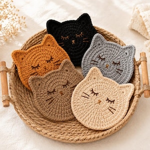 Peut inclure: Un ensemble de cinq dessous de verre ronds en crochet en forme de chat, de couleurs noir, jaune moutarde, gris clair, beige et crème. Chaque dessous de verre présente des yeux fermés, un petit nez et des moustaches brodés, dans un panier en osier.