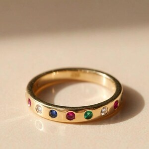 Puede incluir: Un anillo de banda de oro con una serie de piedras preciosas de corte redondo engastadas al ras en colores alternos que incluyen rojo, azul, verde y blanco transparente. La superficie del metal es pulida y lisa, mostrando un diseño circular.