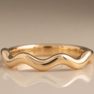 May include: A polished gold ring featuring a continuous, smooth, undulating wave pattern around the entire band. The metal has a high-shine, reflective finish that catches the light, creating a fluid, rhythmic shape that circles the finger.