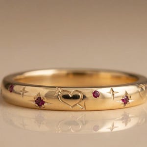 Puede incluir: Un anillo de banda de oro con motivos grabados de corazones y flechas, intercalados con pequeños rubíes redondos de color rosa intenso engastados en tallas en forma de estrella. La superficie de metal pulido refleja la luz y resalta los detalles.