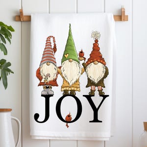 May include: A white kitchen towel with three gnomes standing on the word "JOY" in black. The gnomes are wearing red, green, and brown hats and have white beards. A small red cardinal is perched on the word "JOY".