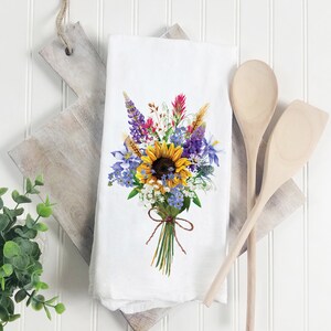 May include: White kitchen towel with a colorful floral bouquet design. The bouquet features a large yellow sunflower, purple lupine, blue forget-me-nots, and red Indian paintbrush.