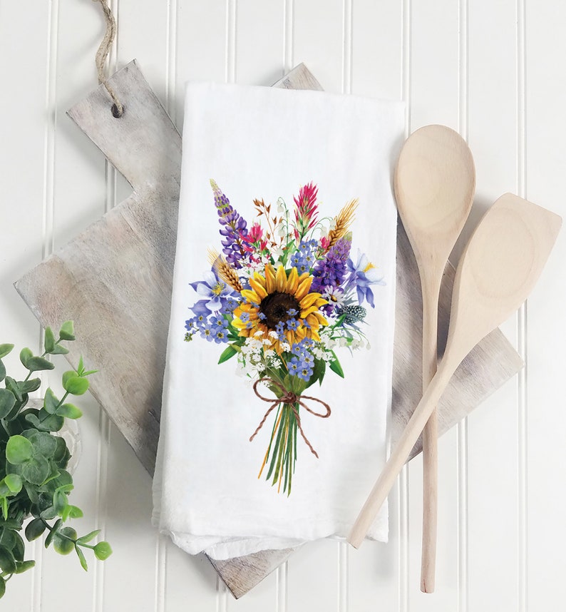 May include: White kitchen towel with a colorful floral bouquet design. The bouquet features a large yellow sunflower, purple lupine, blue forget-me-nots, and red Indian paintbrush.