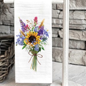 May include: White bath towel with a watercolor floral bouquet design featuring sunflowers, bluebells, and other wildflowers. The towel is 19x28 inches and is displayed on a white towel rack.