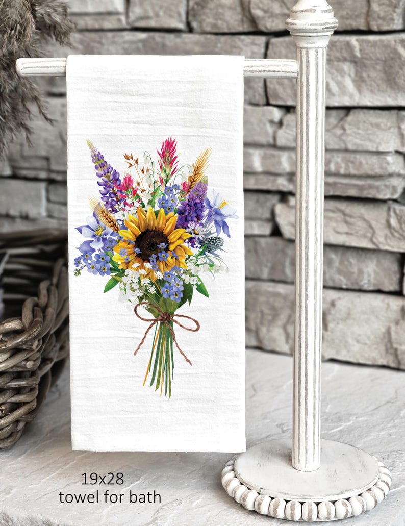May include: White bath towel with a watercolor floral bouquet design featuring sunflowers, bluebells, and other wildflowers. The towel is 19x28 inches and is displayed on a white towel rack.