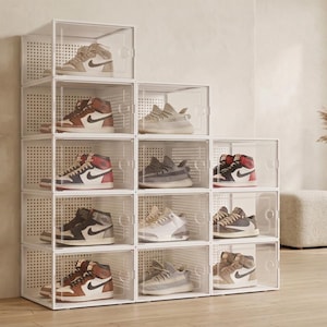 May include: A modular storage system consisting of twelve clear, rectangular plastic shoe boxes stacked in a stepped arrangement. Each box features a transparent front door and a white perforated back panel, housing various athletic sneakers.