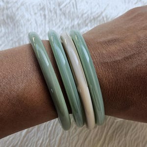Peut inclure: Un ensemble de quatre bracelets joncs lisses et arrondis portés au poignet. Trois des bracelets sont d'une couleur vert sauge unie, tandis que le bracelet central est d'une couleur crème unie. Chaque bracelet a une finition brillante et lisse.