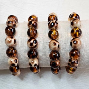Peut inclure: Quatre bracelets de perles disposés en rangée, chacun présentant un motif répétitif de perles rondes. Les perles alternent entre des sphères translucides brun ambre foncé et des sphères crème opaques avec des taches léopard brun foncé.