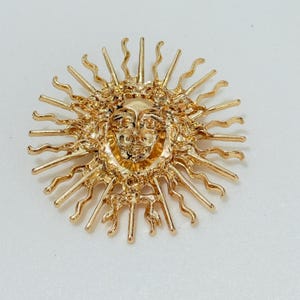 Könnte beinhalten: Eine goldfarbene Metallbrosche in Form einer Sonne mit einem menschlichen Gesicht in der Mitte. Das Design zeigt abwechselnd gerade und gewellte Strahlen, die vom Gesicht nach außen ragen und ein kreisförmiges Strahlenmuster bilden.