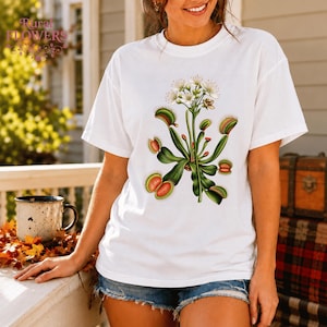 Può includere: Una t-shirt bianca a maniche corte con un'illustrazione botanica di una pianta venere acchiappamosche con foglie verdi, trappole bordate di rosso e piccoli fiori bianchi centrati sulla parte anteriore del capo.