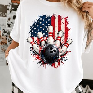 May include: A white t-shirt featuring a graphic print of a black bowling ball striking five white bowling pins with red stripes. Behind the bowling equipment is a stylized, distressed American flag design with red, white, and blue colors.