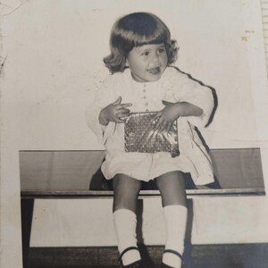 Pode incluir: Uma fotografia vintage a preto e branco de uma criança pequena com cabelo pelos ombros, vestindo um vestido claro de manga comprida com botões, meias brancas e sapatos escuros. A criança sorri e segura um pequeno presente retangular.