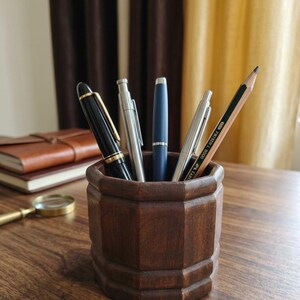 May include: A dark brown wooden pencil holder with an octagonal shape containing a black fountain pen, a silver mechanical pencil, a matte blue ballpoint pen, a thin silver pen, and a wooden pencil with a black and gold striped design.