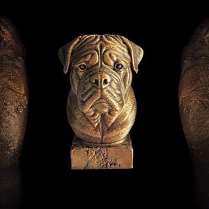 Puede incluir: Una escultura de perro de color bronce, que presenta un busto detallado de una raza de perro con una cara arrugada y orejas caídas. La escultura está montada sobre una base rectangular, sobre un fondo negro. La expresión del perro es seria.