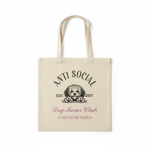 Op de afbeelding: Crèmekleurige draagtas met de woorden "ANTI SOCIAL" boven een hondenillustratie. Daaronder staan "Dog Moms Club" en "PAWS BEFORE PEOPLE" in roze letters. De tas heeft twee crèmekleurige handvatten.
