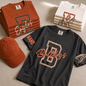 May include: A collection of short-sleeve t-shirts and a baseball cap. The shirts feature collegiate-style lettering with the words Eagles, Mascot, and Bulldogs. One shirt displays the word NAME on the sleeve and a pennant graphic with MASCOT.