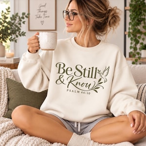 May include: A cream-coloured crewneck sweatshirt featuring dark green text that reads Be Still & Know with the reference Psalm 46:10. The design includes a graphic of a dove holding an olive branch, positioned to the right of the text.