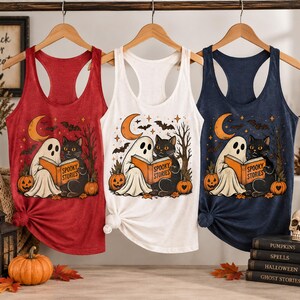 May include: Three racerback tank tops in red, white, and navy blue, each featuring a graphic print of a ghost and a black cat reading a book titled Spooky Stories, with a crescent moon, bats, and pumpkins in the background of the illustration.