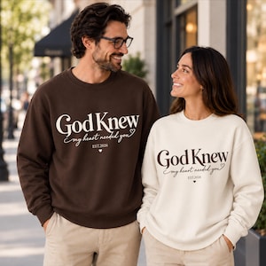May include: A dark brown sweatshirt and a cream-coloured sweatshirt, both featuring the text God Knew my heart needed you EST.2026 with a small heart icon. The dark brown sweatshirt has white text, while the cream sweatshirt has dark brown text.