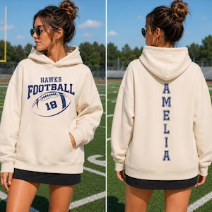 May include: A cream-coloured hooded sweatshirt featuring a dark blue graphic on the front with the text HAWKS FOOTBALL and the number 18 inside a football illustration. The back displays the name AMELIA printed vertically in dark blue block letters.
