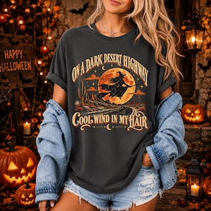 May include: A charcoal grey t-shirt featuring a graphic of a witch flying on a broomstick in front of a large orange moon, with a desert landscape and the text ON A DARK DESERT HIGHWAY COOL WIND IN MY HAIR printed in a vintage-style font.