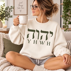 May include: A cream-coloured crewneck sweatshirt featuring dark olive green text on the front. The design includes the Hebrew characters for YHWH positioned above the English letters YHWH in a clean, bold, sans-serif font style.