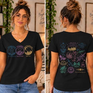 May include: A black V-neck t-shirt featuring three circular graphic designs on the front with the text Terrasen, Velaris, and Lunathion. The back displays a collection of colourful fantasy-themed illustrations and various quotes.