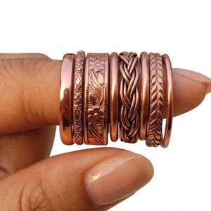 Op de afbeelding: Een set van zeven koperen ringen met diverse texturen en ontwerpen, waaronder een gladde band, een geometrisch patroon, een bloemmotief, een gevlochten stijl en een bladvormig patroon, allemaal samen op een rij aan een vinger.